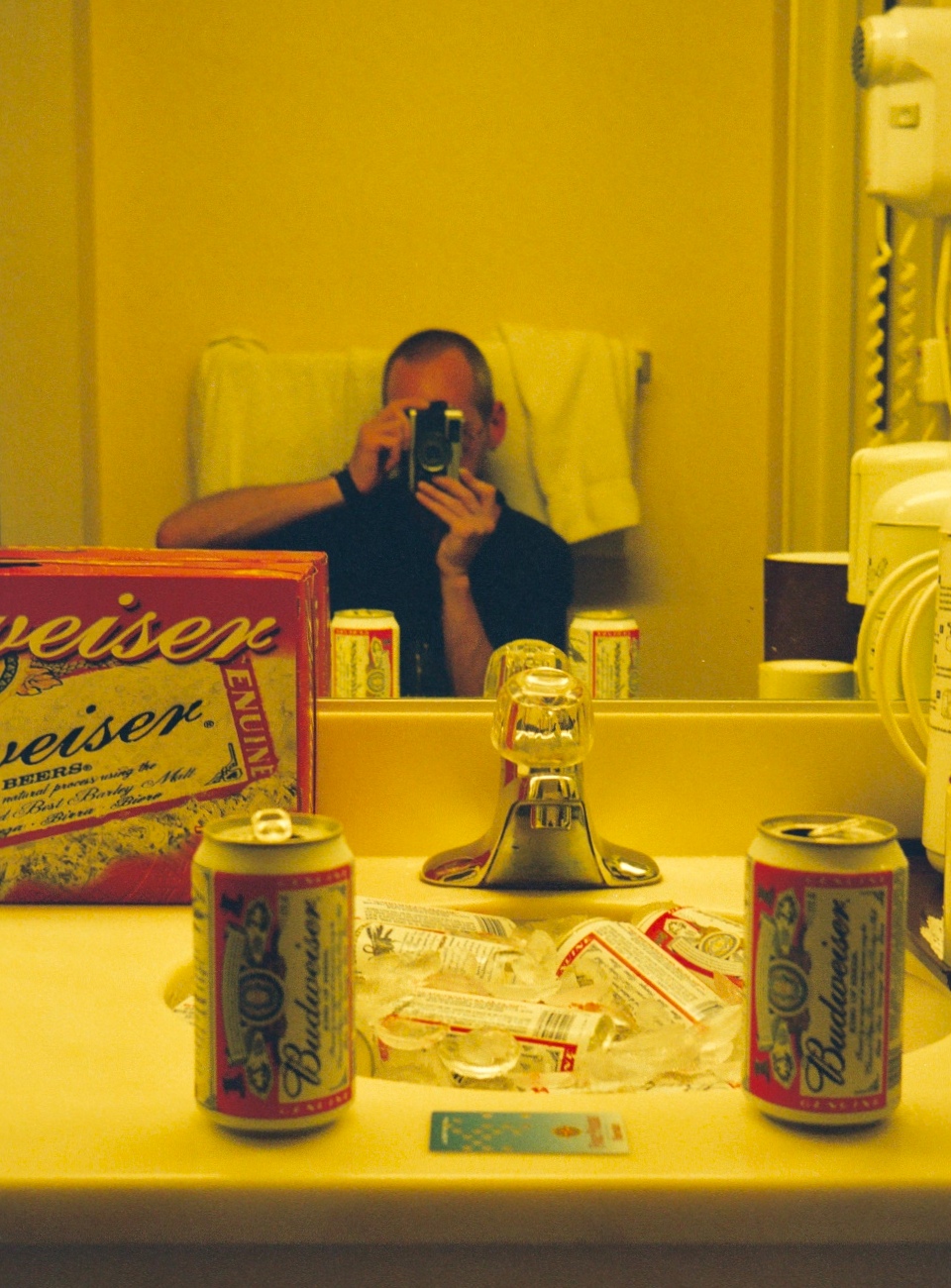 Self-portrait of Simon in a hotel room mirror, with the sink full of ice and cans of beer.